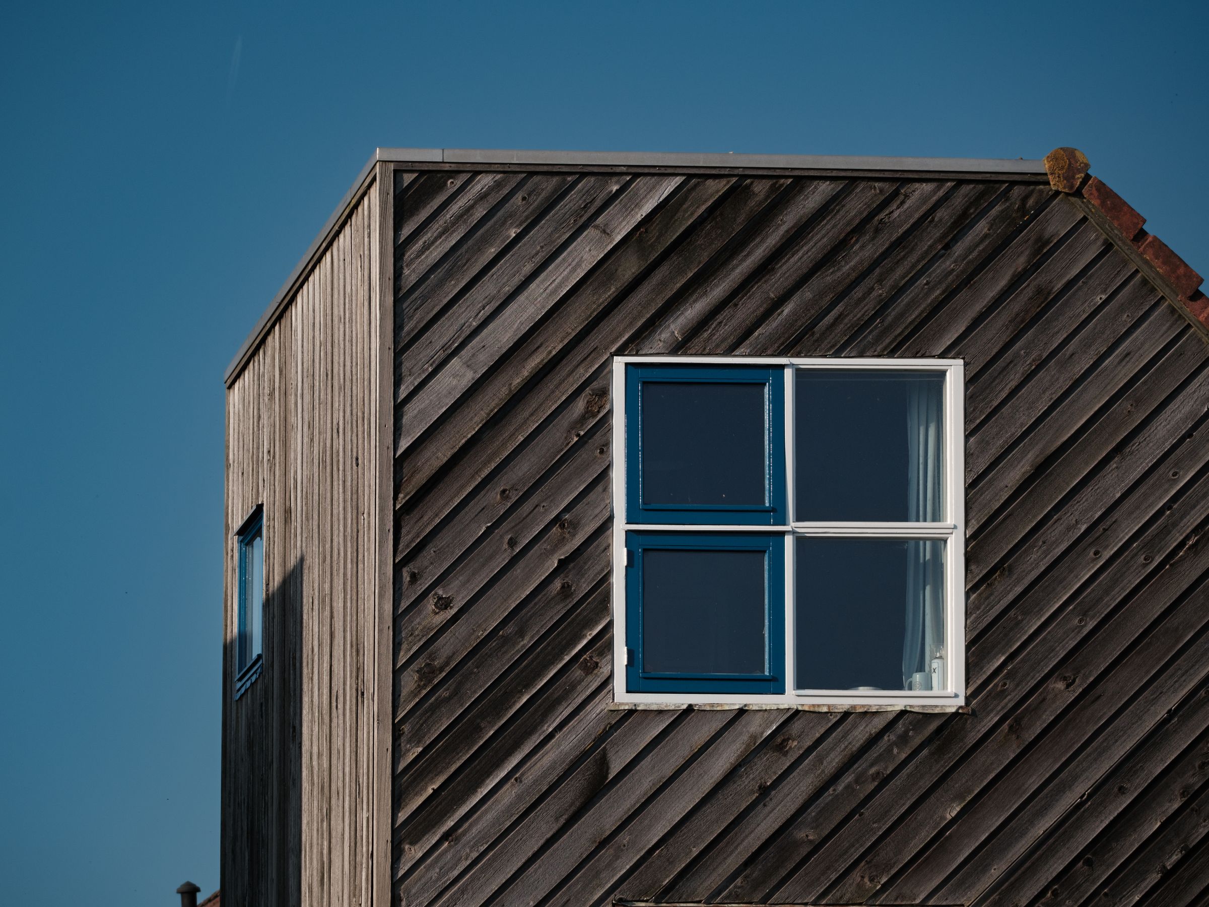 Dark timber-clad house corner with a large window against a clear sky